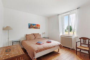Une chambre à coucher comprenant un lit, une chaise, une table, un radiateur, une fenêtre avec des rideaux et un tableau accroché au mur.