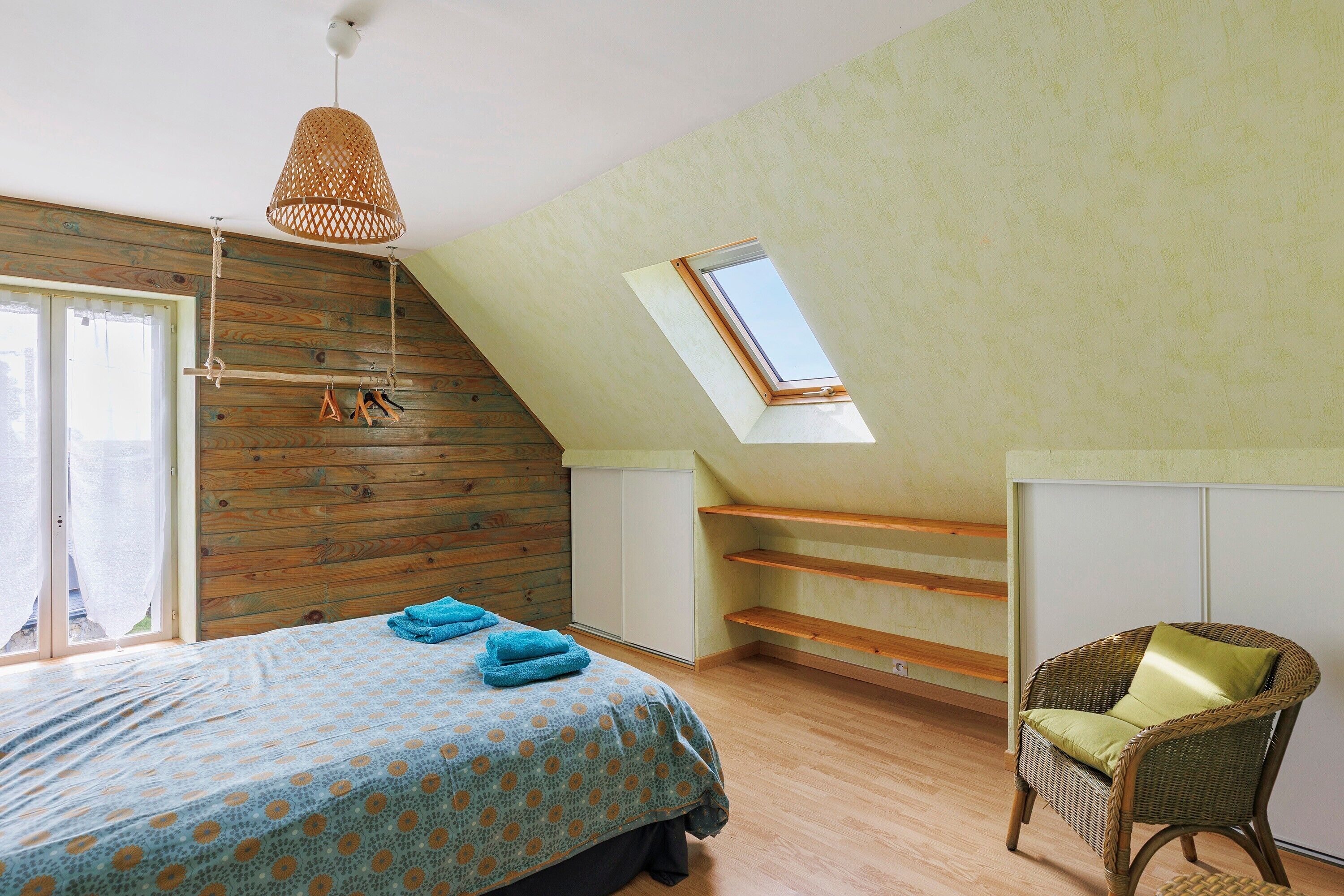 Une chambre avec un plafond en pente, un lit recouvert d’une couvre-lit à motifs, une chaise en osier et un puits de lumière.