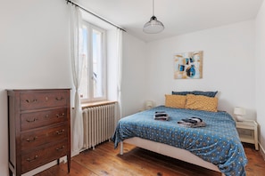 Un dormitorio con cama, cómoda, radiador, ventana con cortinas, un cuadro en la pared y una lámpara colgante.