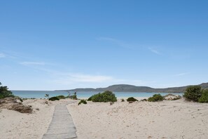 En sandstrand med en träpromenad som leder ner till vattnet, omgiven av gles vegetation och avlägsna kullar.