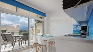 Une cuisine moderne dotée d’une machine à café intégrée, d’un coin repas avec des chaises et une table blanches, et de grandes fenêtres offrant une vue sur l’extérieur.