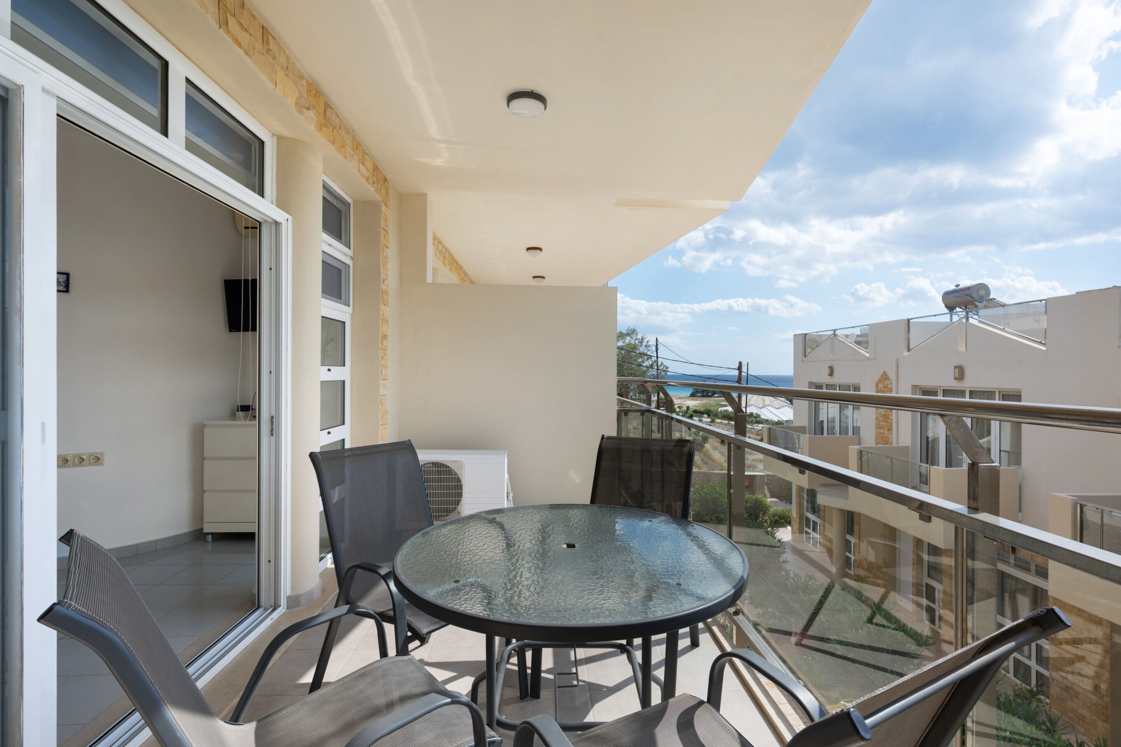 Un balcon avec une table et des chaises en verre, un meuble blanc et une vue sur la mer.