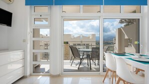 Une cuisine moderne équipée d’un téléviseur intégré, un coin repas avec une table et des chaises, et une porte-fenêtre coulissante donnant sur une terrasse extérieure avec vue sur les bâtiments et les montagnes.