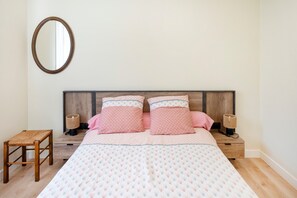A bedroom with a bed, two bedside tables, a small stool, a wall-mounted mirror, and a wooden headboard. - “Garden Level” Apartment (Hendaye)