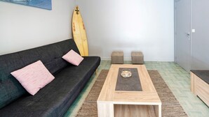 Una stanza con un divano grigio scuro, due cuscini rosa, un tavolino in legno con un vassoio decorativo, un surf che appoggia al muro e una porta sulla destra.