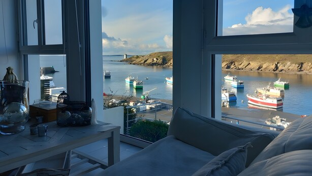 Ein Zimmer mit großem Fenster mit Blick auf einen Hafen, in dem Boote liegen, einen Leuchtturm und eine felsige Küste.