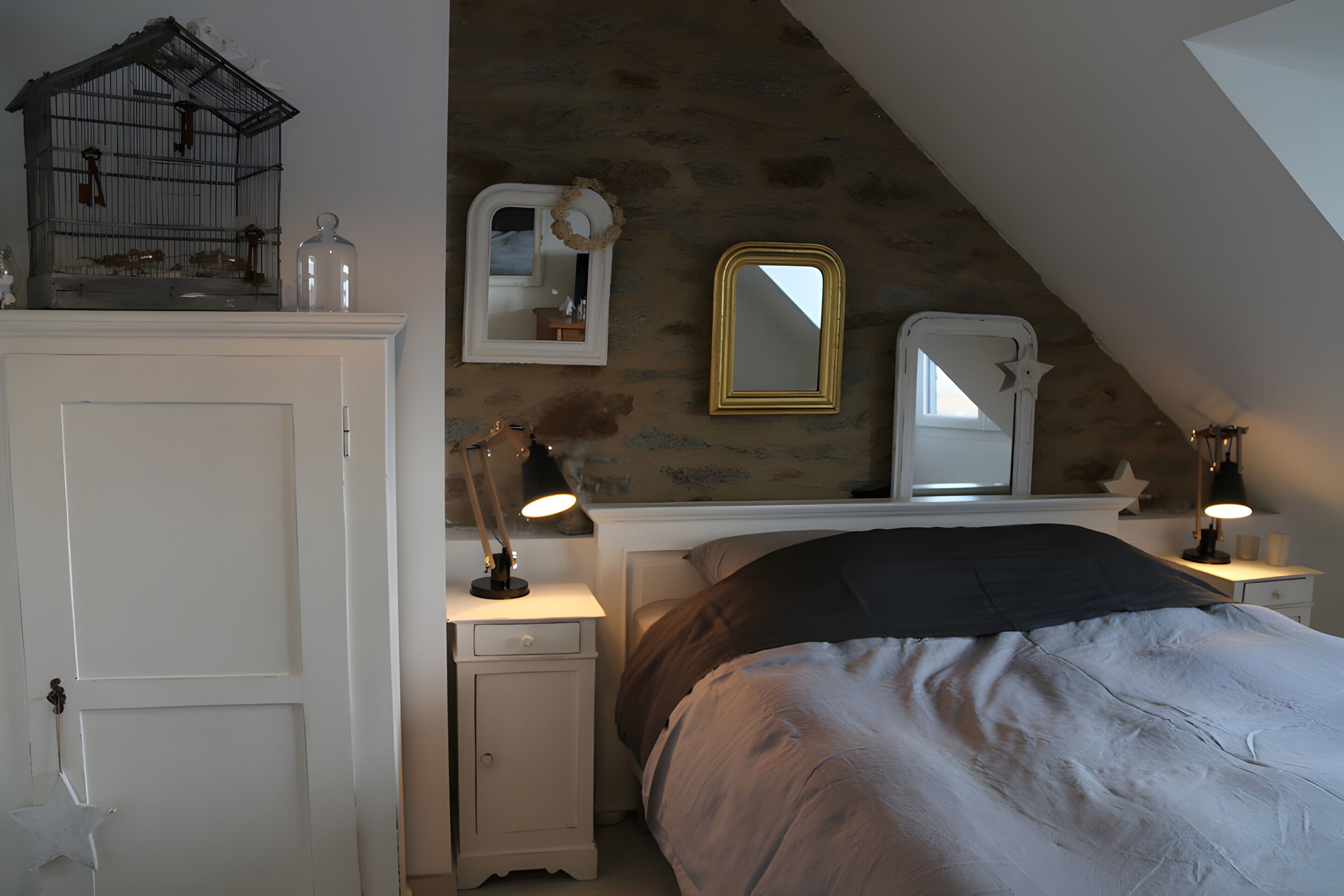 A bedroom with a sloped ceiling, a bed with a dark headboard, a dresser with a birdcage, a mirror, and a nightstand with a lamp.