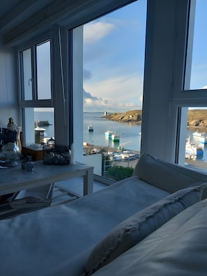A room with a large window overlooking a harbor, a table with decorative items, and a bed with pillows. - Harborfront Seaside House with Port View (Le Conquet)