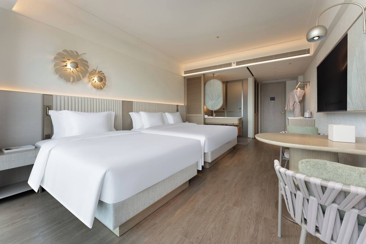 A modern hotel room with two beds, a sitting area, a vanity with a mirror, and a television.