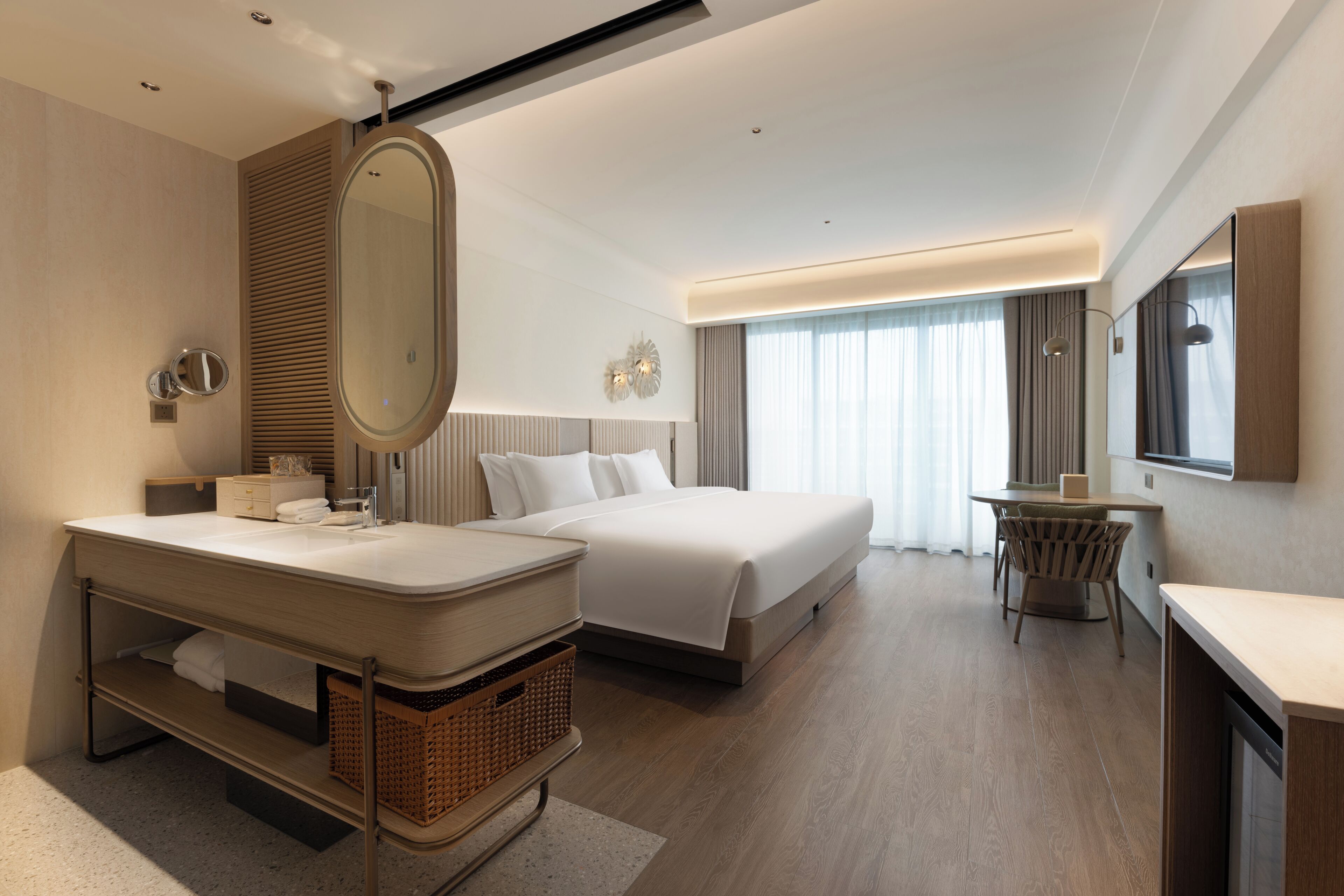 A modern hotel room with a large bed, a separate sitting area, a vanity with a mirror, and a large window with curtains.