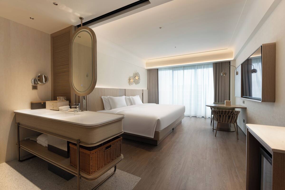 A modern hotel room with a large bed, a separate sitting area, a vanity with a mirror, and a large window with curtains.