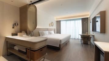 A modern hotel room with a large bed, a separate sitting area, a vanity with a mirror, and a large window with curtains.