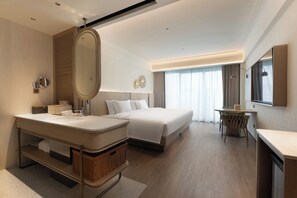 A modern hotel room with a large bed, a separate sitting area, a vanity with a mirror, and a large window with curtains.