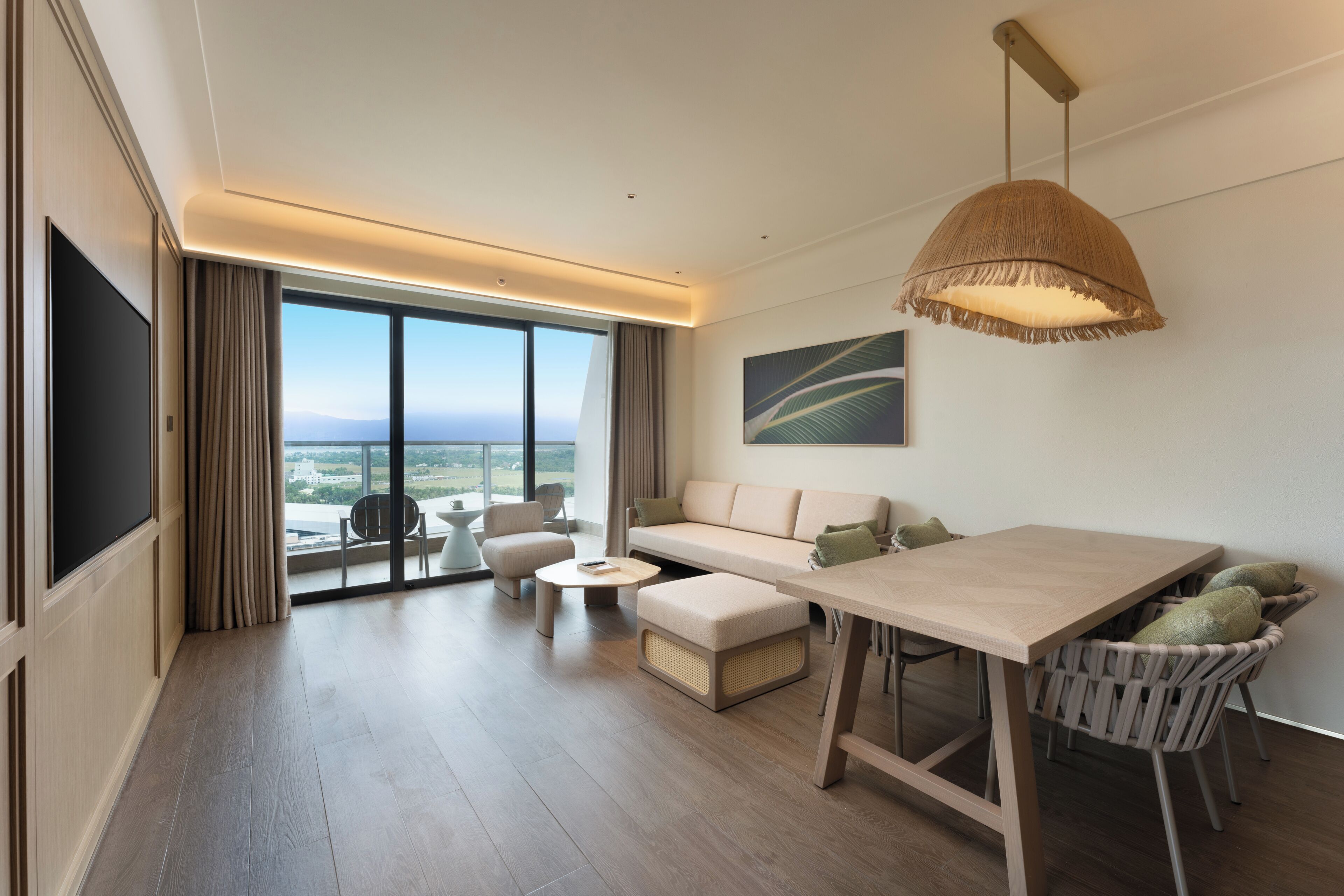 A modern hotel room with a large window, a sofa, a dining table, and a TV.