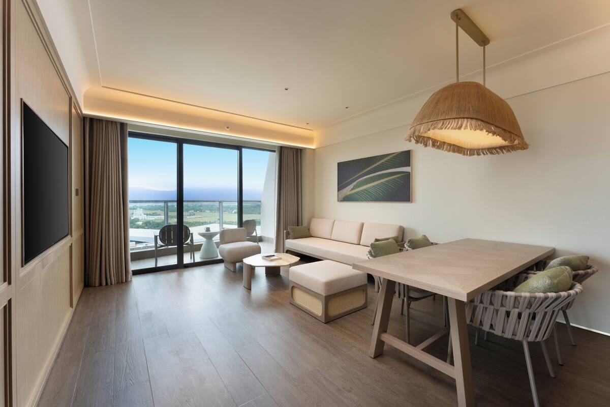 A modern hotel room with a large window, a sofa, a dining table, and a TV.