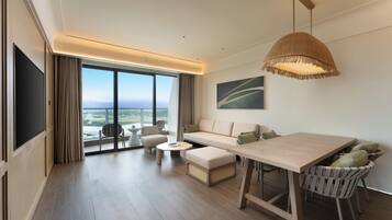 A modern hotel room with a large window, a sofa, a dining table, and a TV.