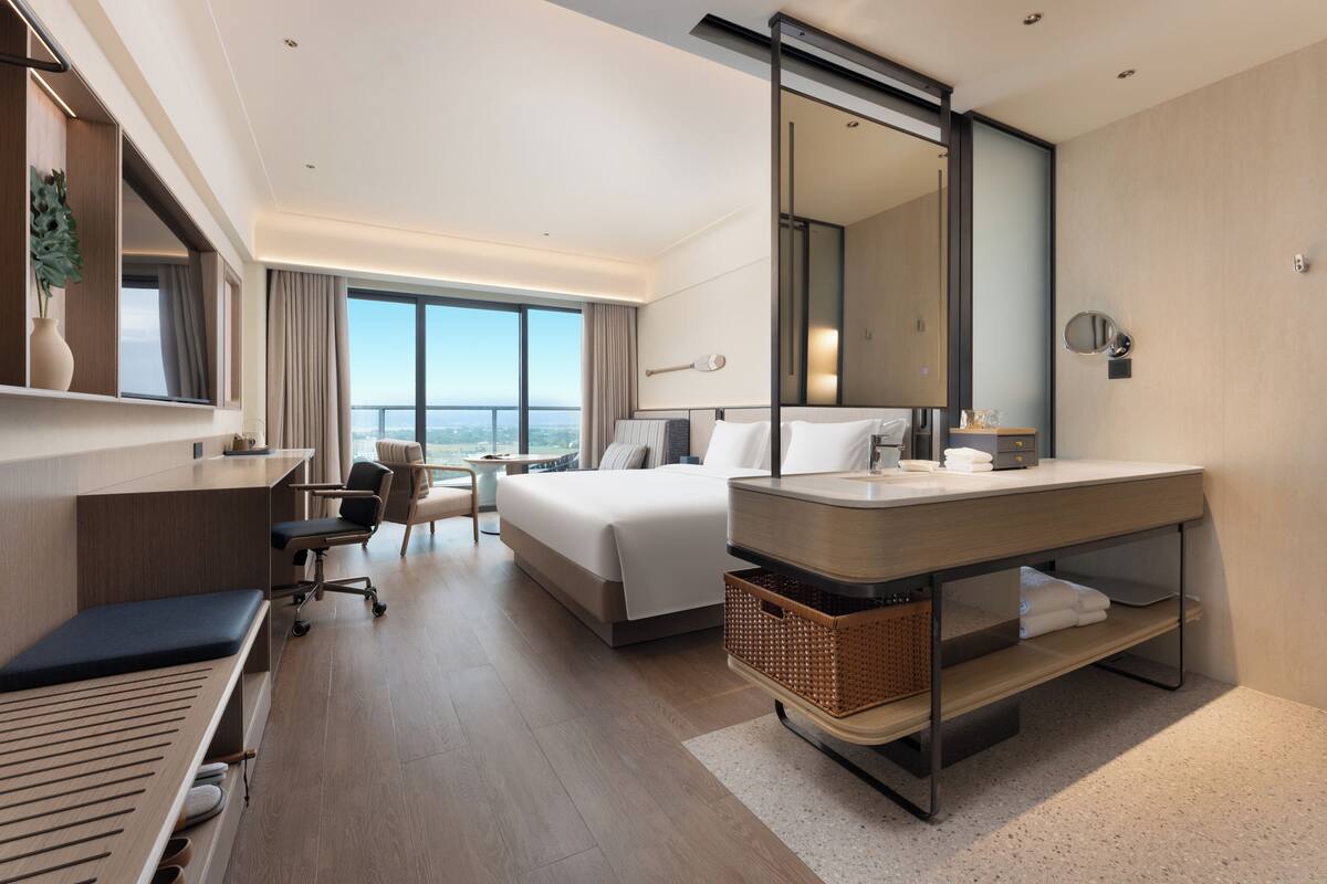 A modern hotel room with a bed, desk, chair, and large windows offering a city view.