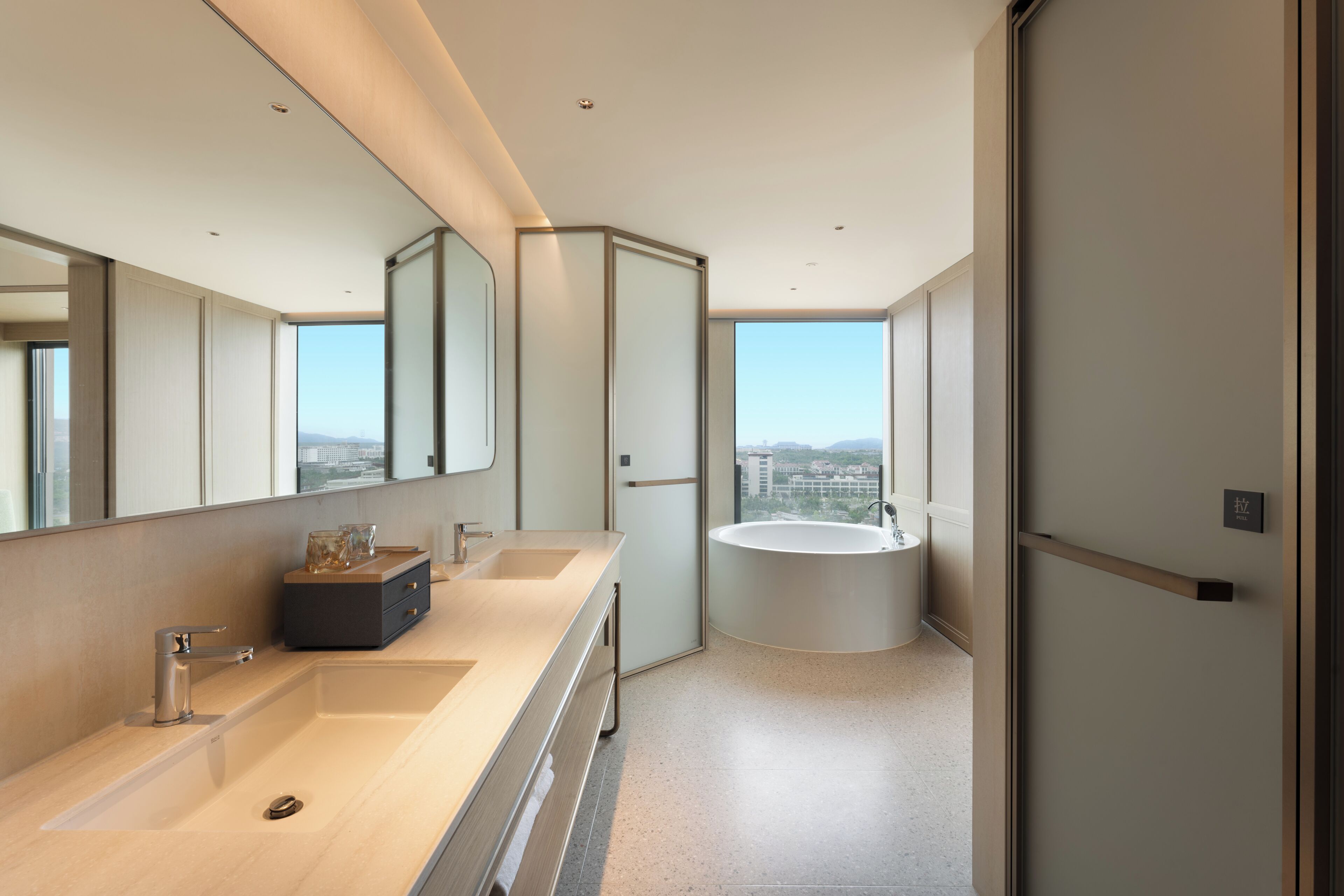 A modern bathroom with a large mirror, two sinks, a bathtub, and a city view.