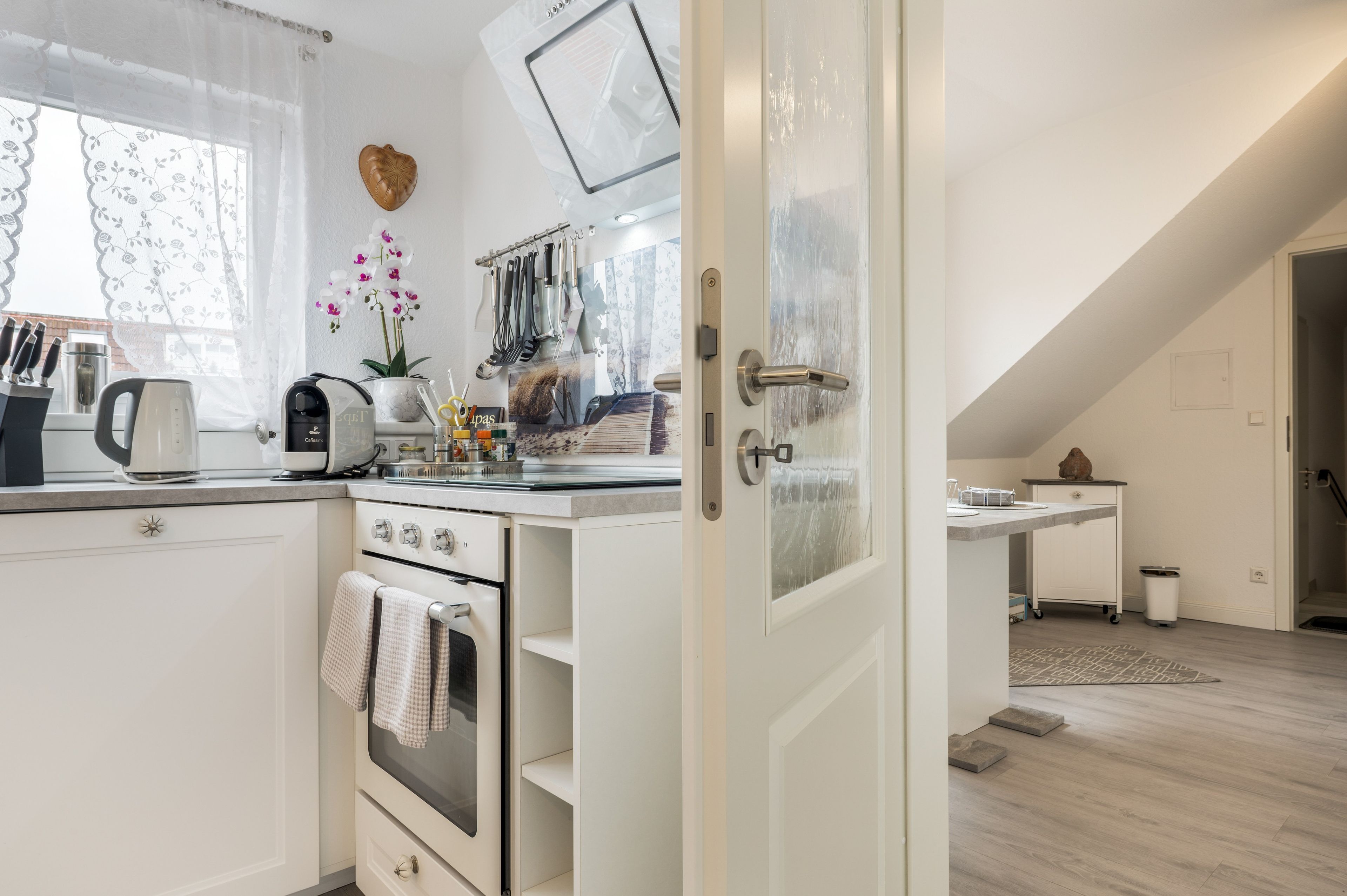 A modern kitchen with white cabinetry, a coffee machine, and a window with lace curtains.