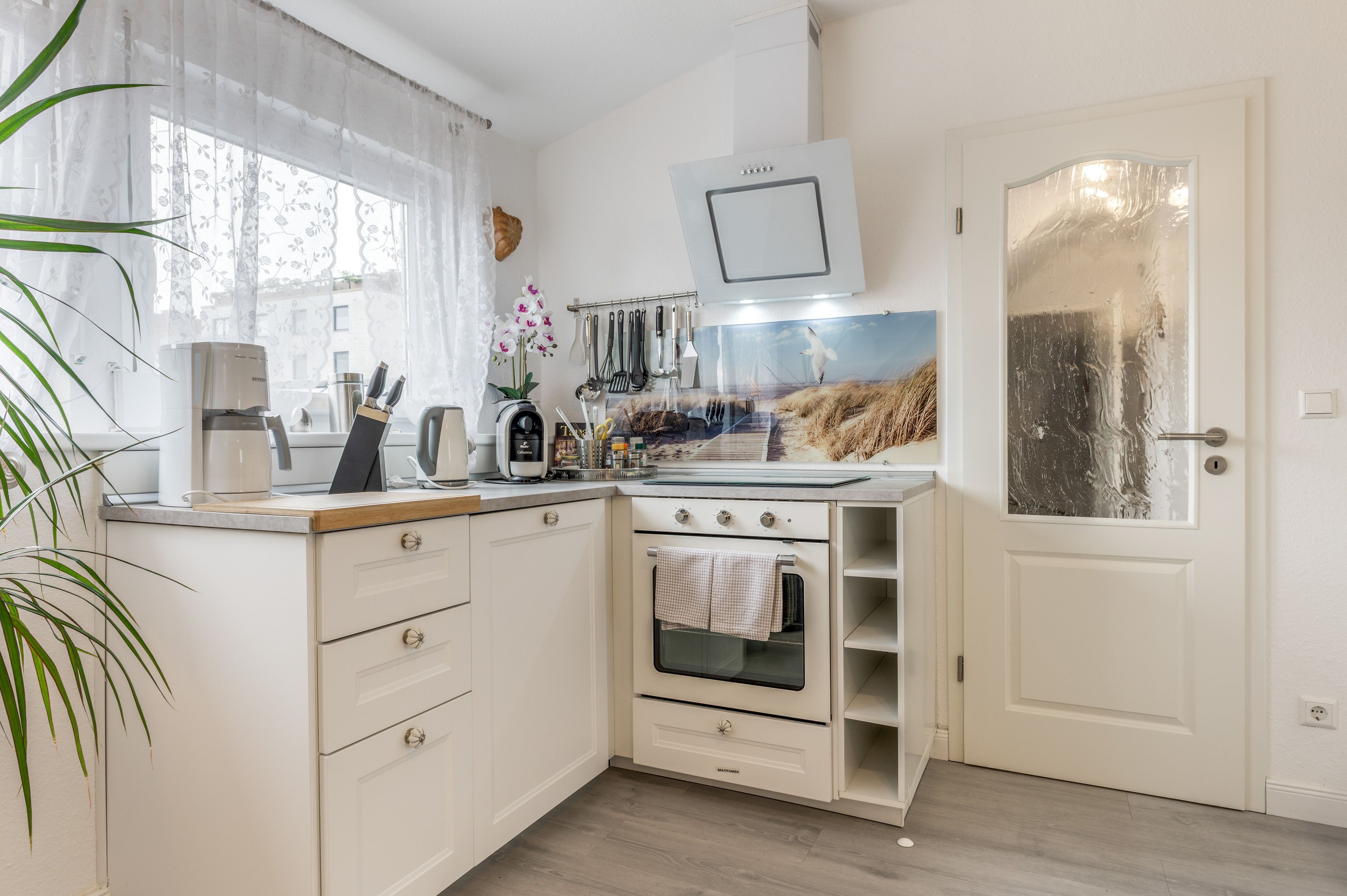 Una cocina compacta con horno integrado, cafetera y ventana con cortinas ligeras.