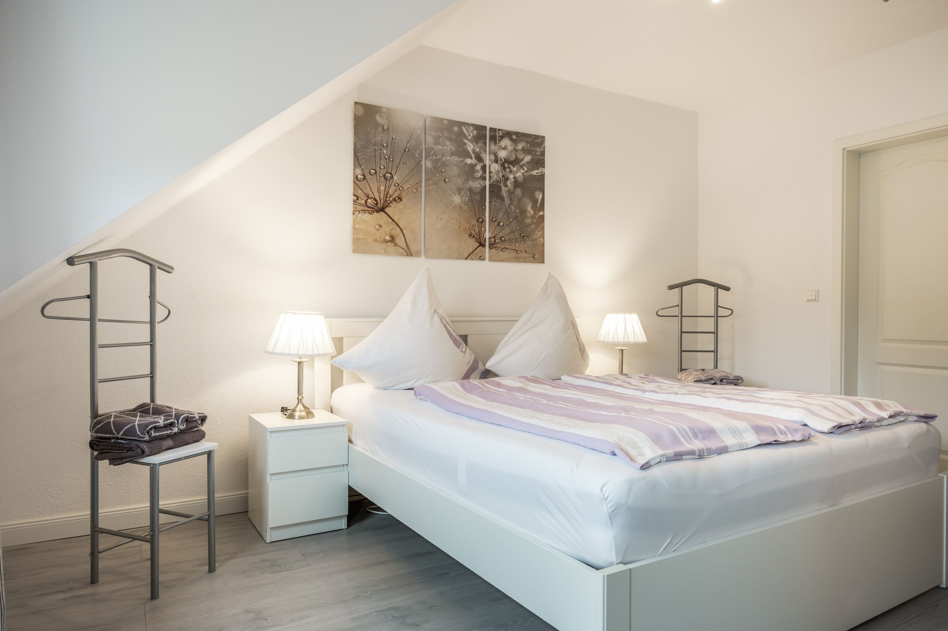 Un dormitorio con una cama, dos mesitas de noche, una lámpara de pared, un perchero y un cuadro.