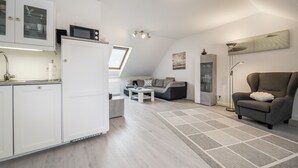 A modern kitchen with white cabinets, a microwave, and a built-in oven. There is a dining area with a sofa and a coffee table. The room has a sloped ceiling with skylights and a large window. - Apartment “Seepferdchen” with Wi-Fi (Cuxhaven)