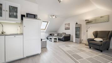Een moderne keuken met witte kastjes, een magnetron en een inbouwoven. Er is een zithoek met een bank en salontafel. De kamer heeft een schuine kap met dakramen en een groot raam.