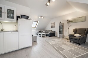 A modern kitchen with white cabinets, a microwave, and a built-in oven. There is a dining area with a sofa and a coffee table. The room has a sloped ceiling with skylights and a large window. - Apartment “Seepferdchen” with Wi-Fi (Cuxhaven)