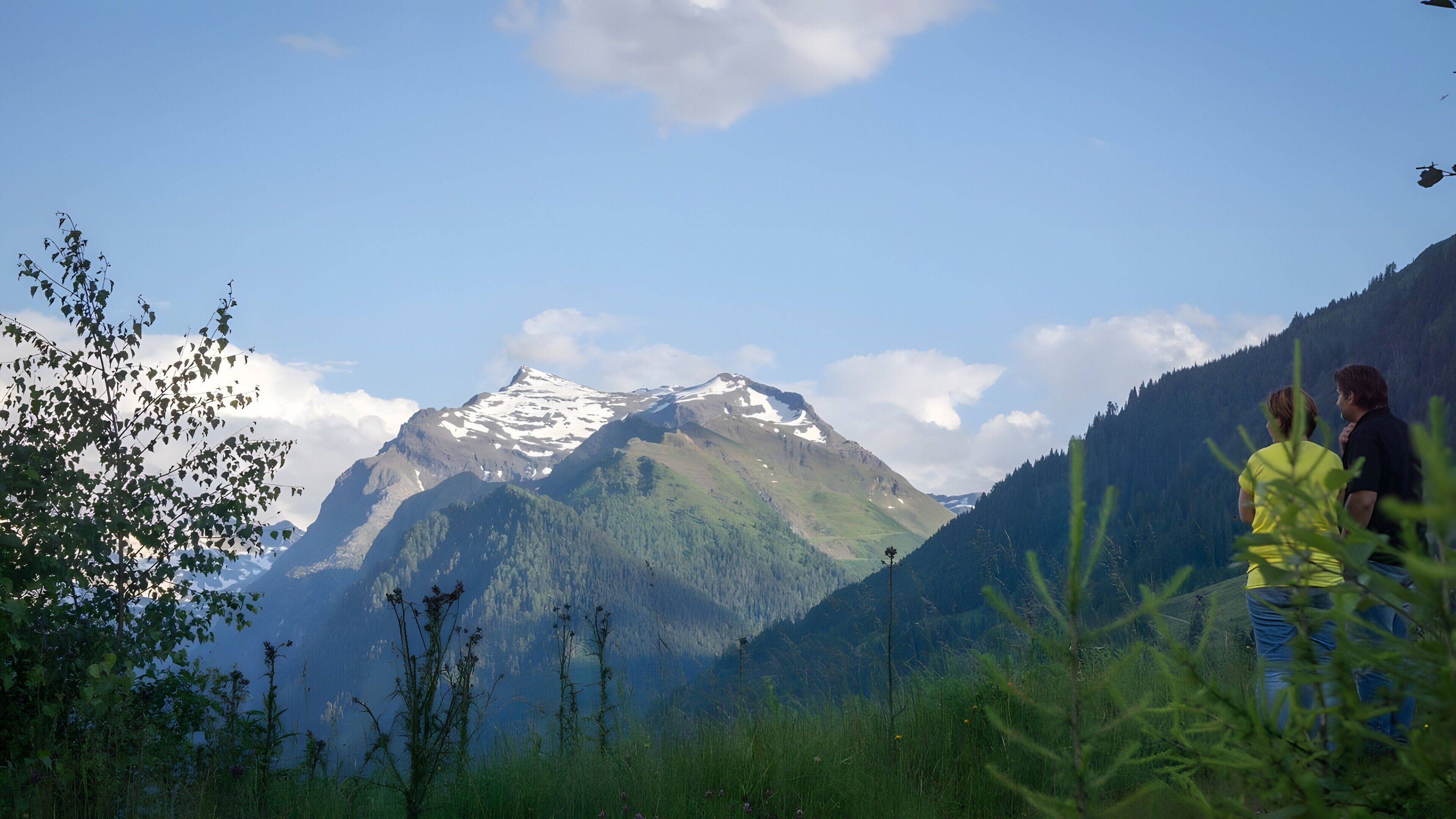 Un paysage de montagne avec un sommet imposant, un couple se tenant au premier plan et un ciel dégagé.