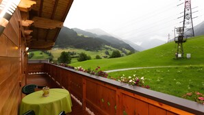 A wooden balcony with a table and chairs, overlooking a green valley with mountains in the distance.