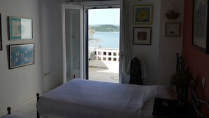 Ein Schlafzimmer mit Bett, Sessel, Tisch und Blick auf ein Gewässer sowie Gebäude.