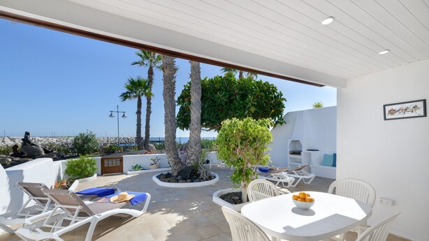Een zonnig terras met witte meubels, een tafel met fruit en uitzicht op zee.