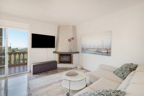 A living room with a fireplace, a sofa, a TV, and a balcony with a view. - Holiday Home 'Between Forest and Sea' with Sea View, Private Terrace and Wi-Fi (Canyelles)