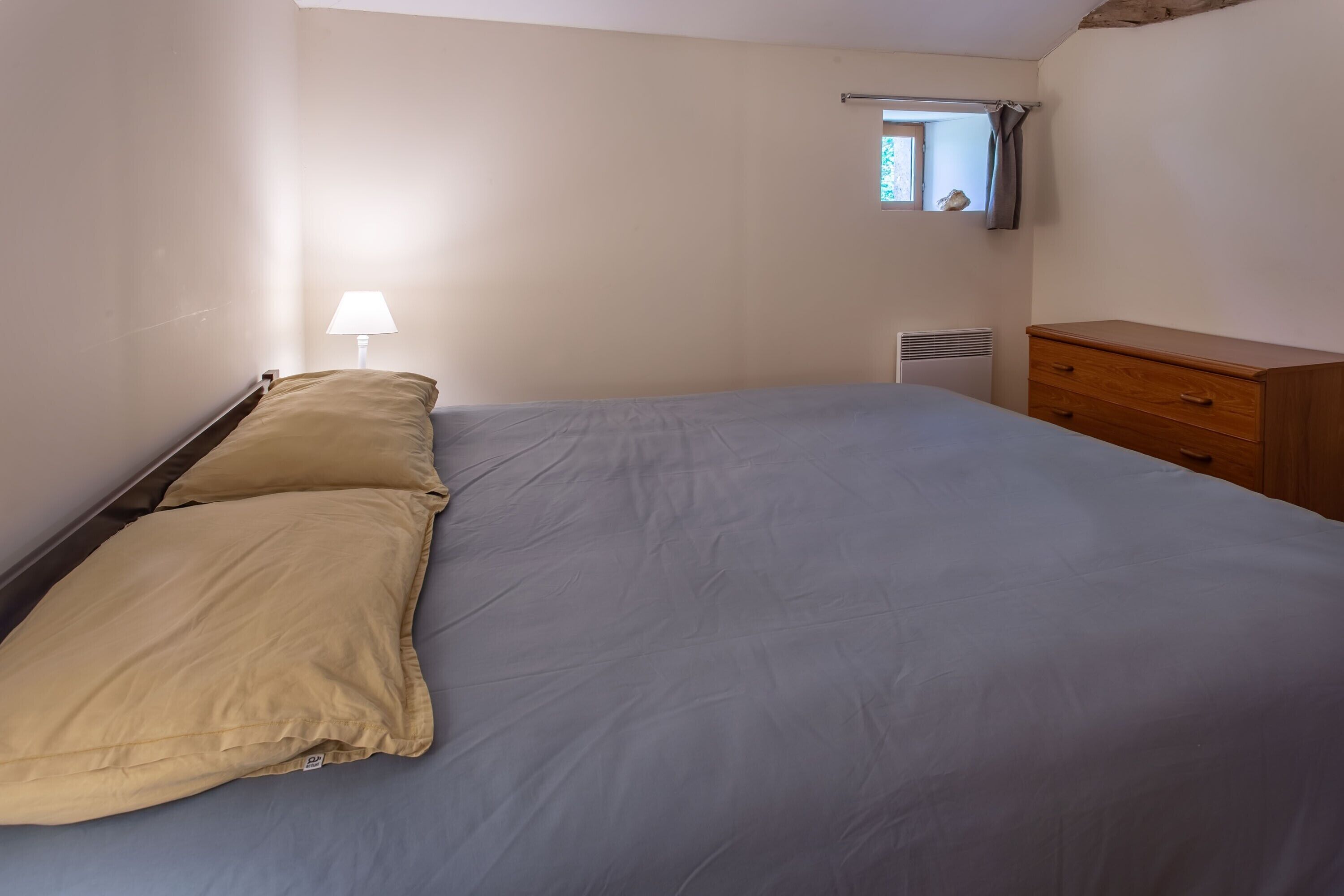 A bedroom with a bed, a nightstand, a lamp, a window with a view, and a dresser.