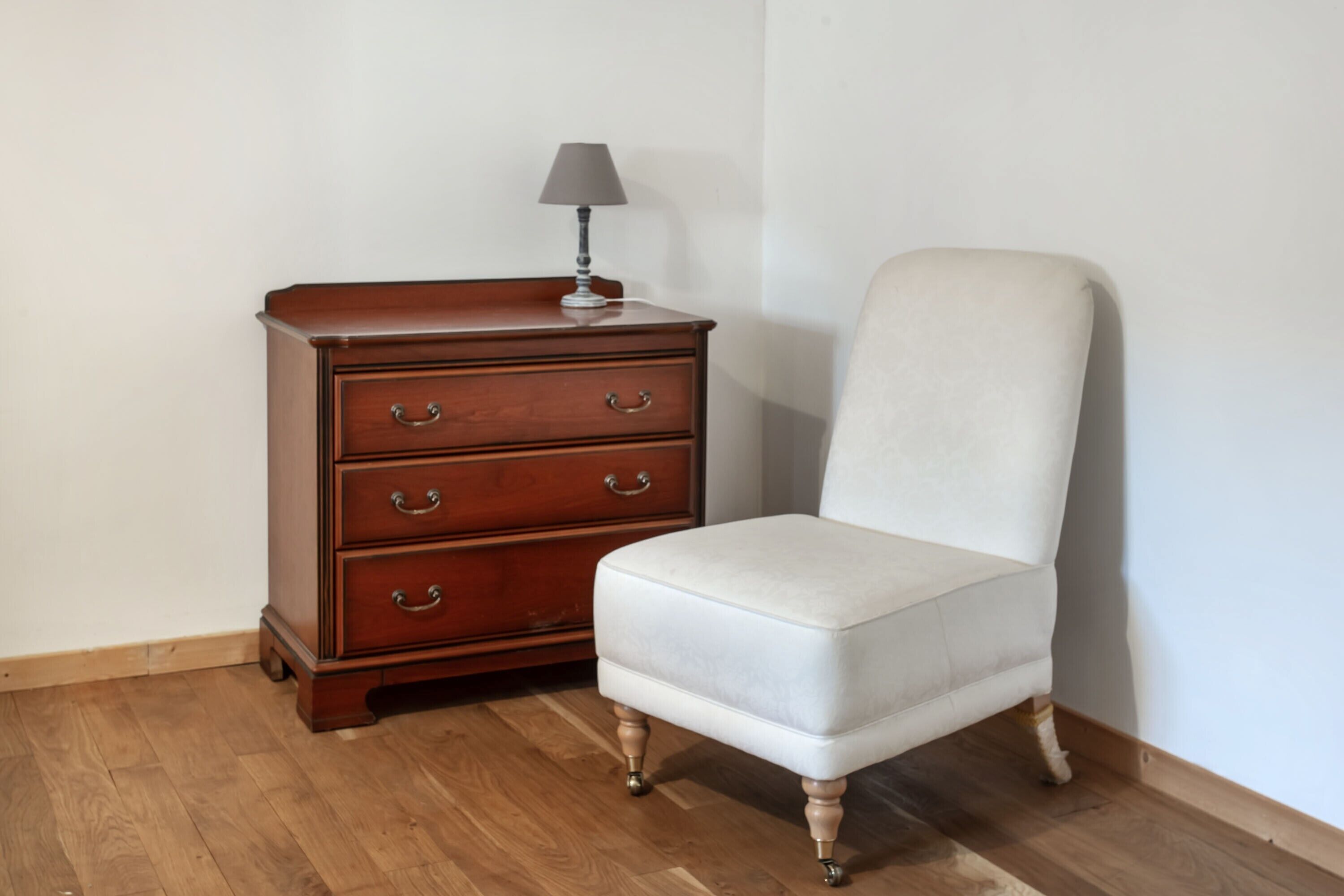 Une commode en bois avec des tiroirs et une lampe, un fauteuil blanc recouvert de tissu et un plancher en bois.