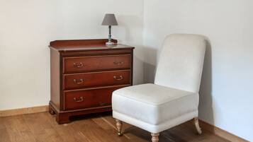 Une commode en bois avec des tiroirs et une lampe, un fauteuil blanc recouvert de tissu et un plancher en bois.