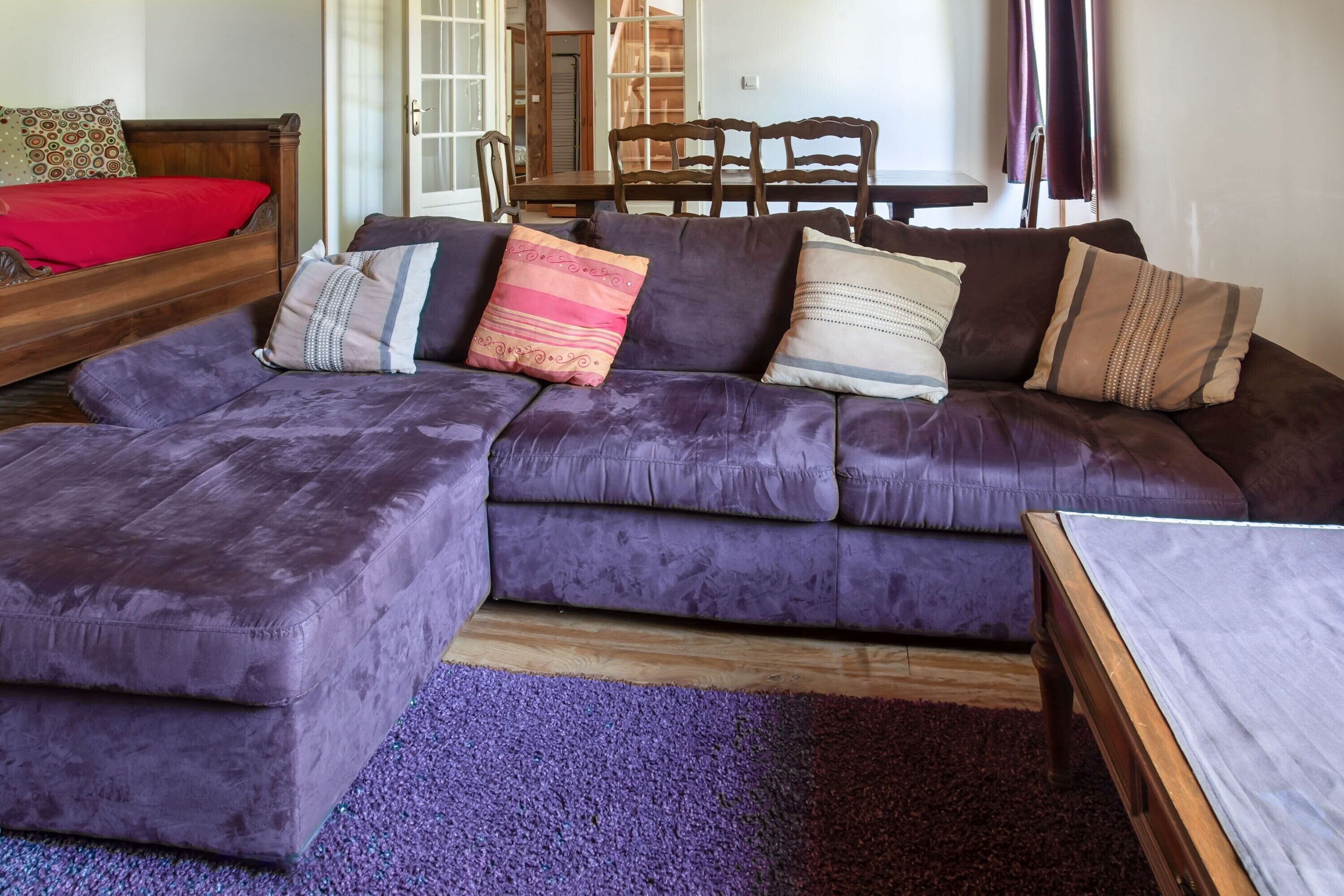 Un salon avec un canapé d’angle violet, une table basse en bois et un lit rouge.