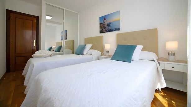 A hotel room with two beds, a wooden door, a large mirror, a painting of a lighthouse, and a nightstand with a lamp. - Apartment 'Playa Arbeyal Garaje' with Wi-Fi (Gijón)