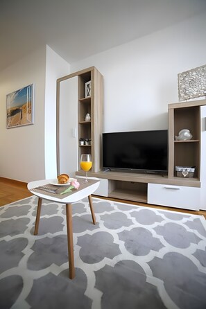Een moderne woonkamer met een flatscreen televisie, een houten kast en een klein tafeltje met een croissant en sinaasappelsap.