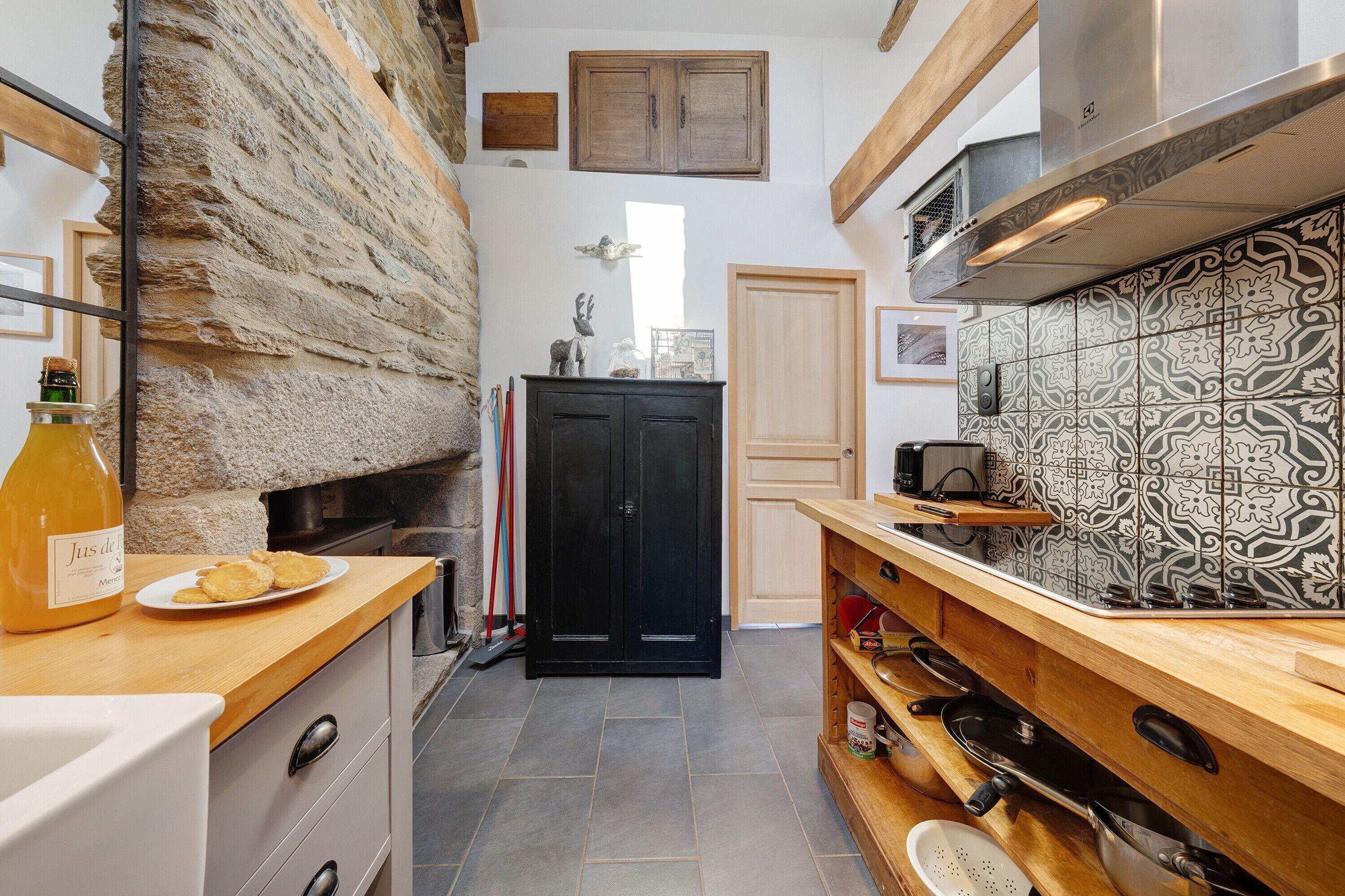 Une cuisine avec des murs en pierre, une porte noire à deux vantaux, un comptoir en bois et un pare-feu en carreaux.