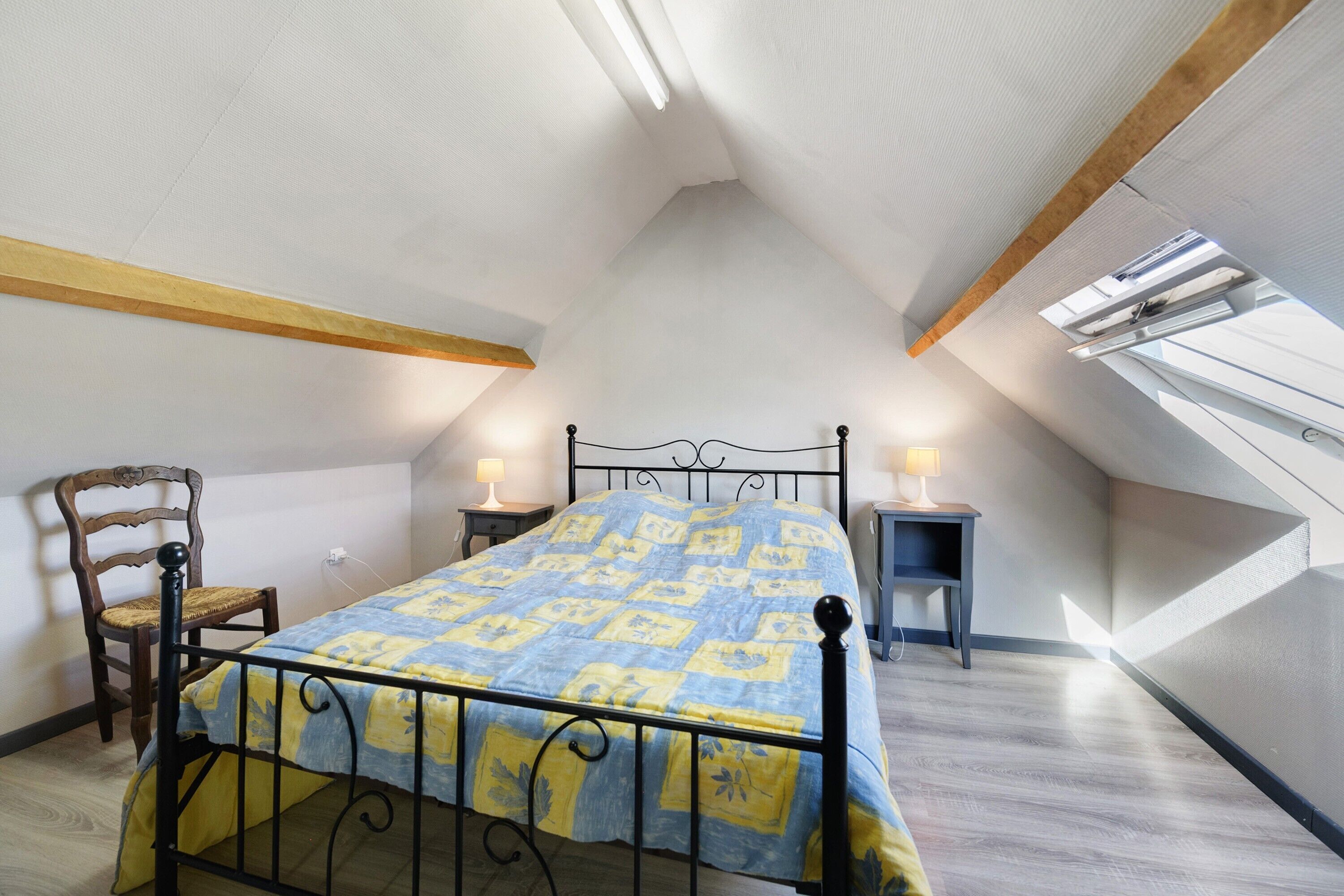 A bedroom with a bed, a chair, two bedside tables, and a sloped ceiling with skylights.