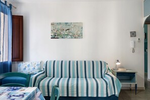 A room with a striped sofa, a small table, a chair, a wall-mounted artwork, and a nightstand with a lamp. - Azzurromare Studio or Rooms Near the Beach, Wi-Fi and Air Conditioning (San Vito Lo Capo)