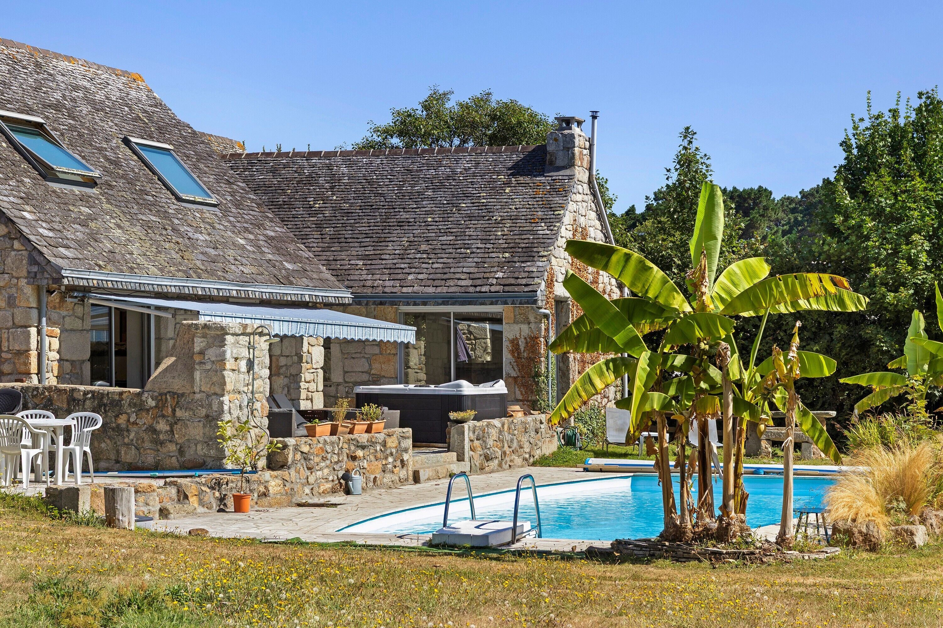 Une maison en pierre avec une piscine, un espace salon extérieur et un jardin planté de bananiers.