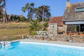 A swimming pool with a stone deck, a stone house with a tiled roof, and a grassy area with trees.