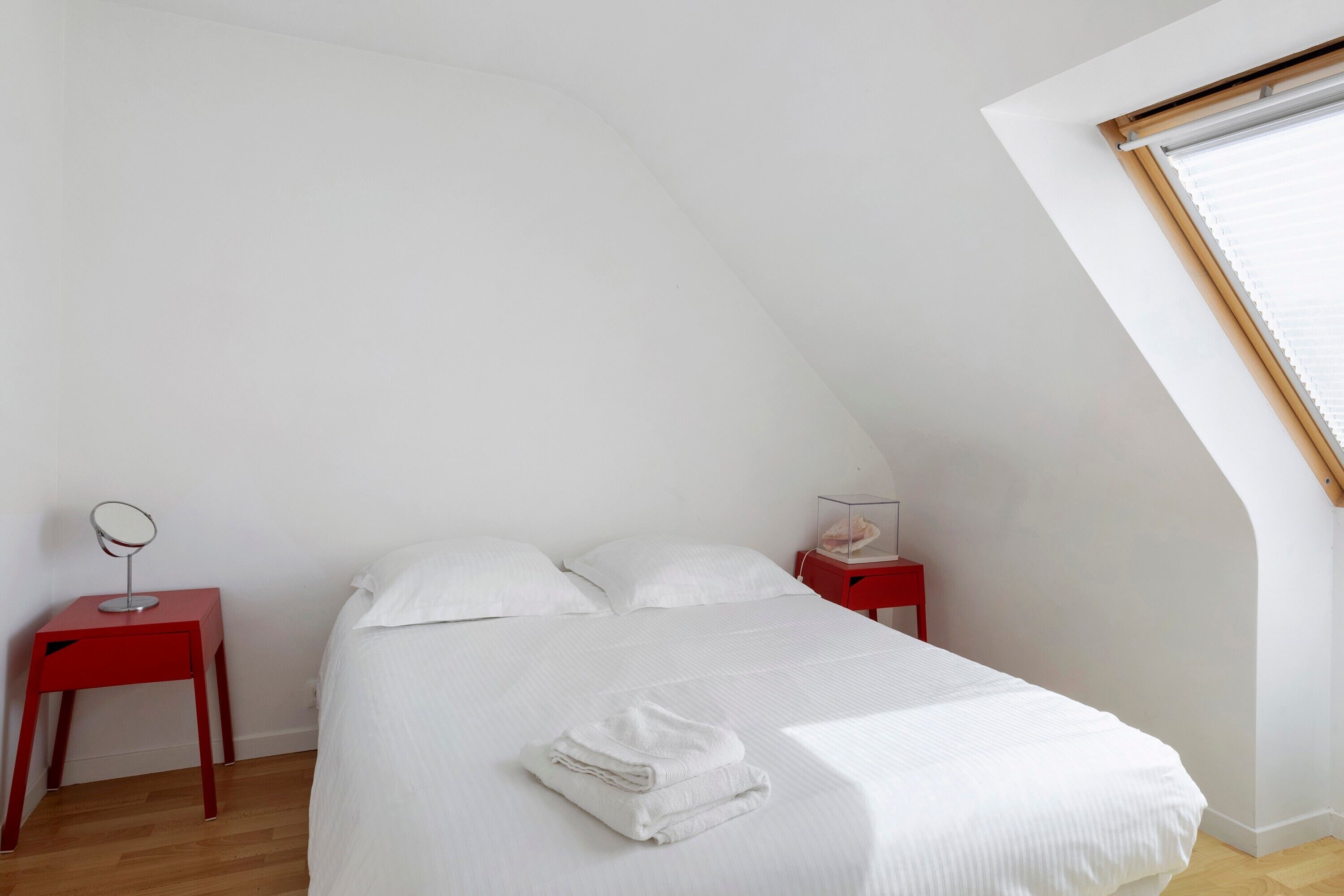 A bedroom with a sloped ceiling, a bed with white linens, two nightstands with red bases, a mirror on one nightstand, and a small decorative item on the other.