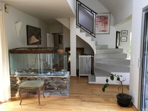 Una habitación amplia con chimenea de piedra, una silla de madera, un acuario y una escalera que sube a un nivel superior.