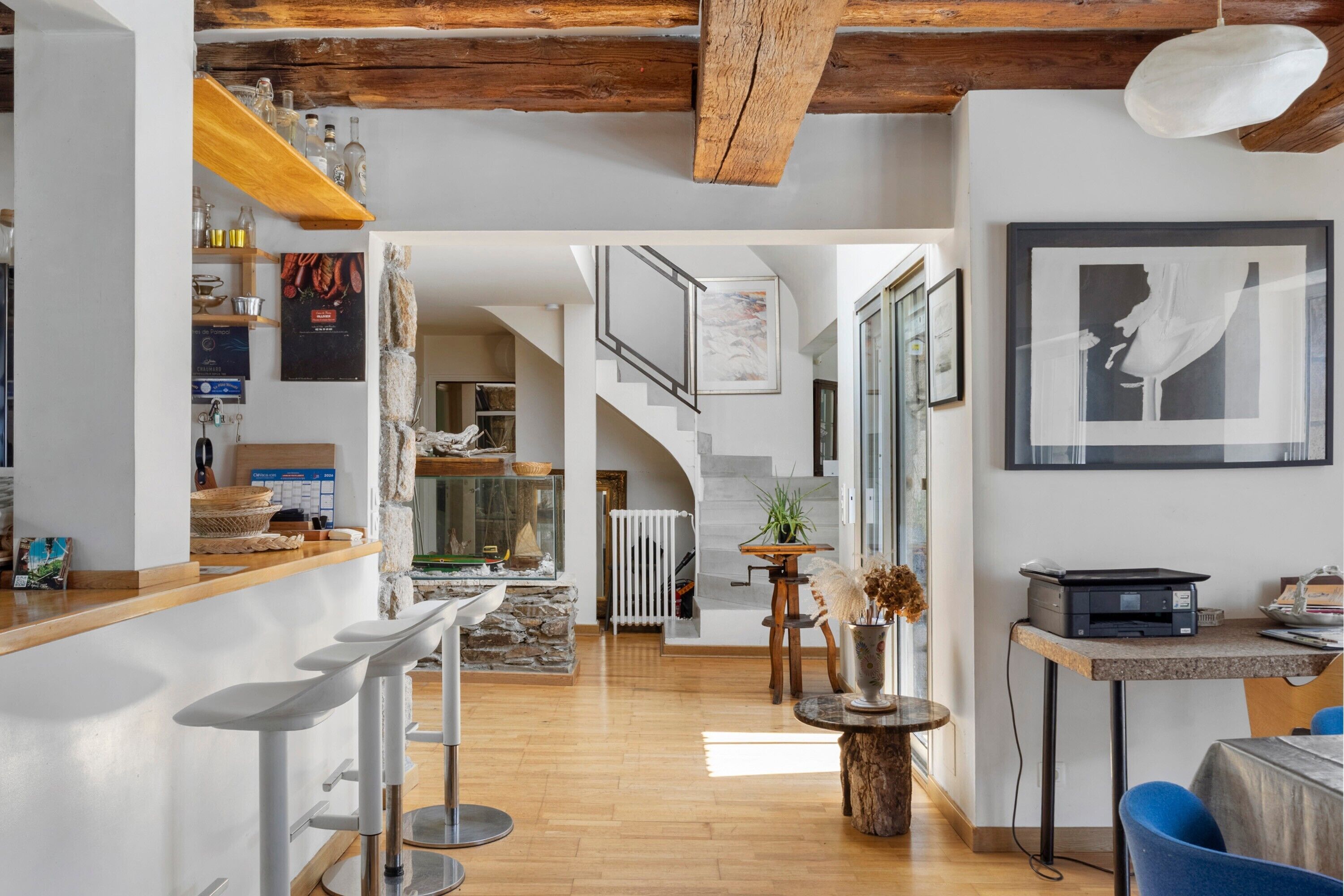 A modern interior with a wooden ceiling, a kitchen area with bar stools, a dining table, and a staircase leading to an upper level.