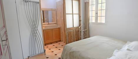 A bedroom with a bed, a wooden wardrobe, a bathroom with a mirror, and a window.