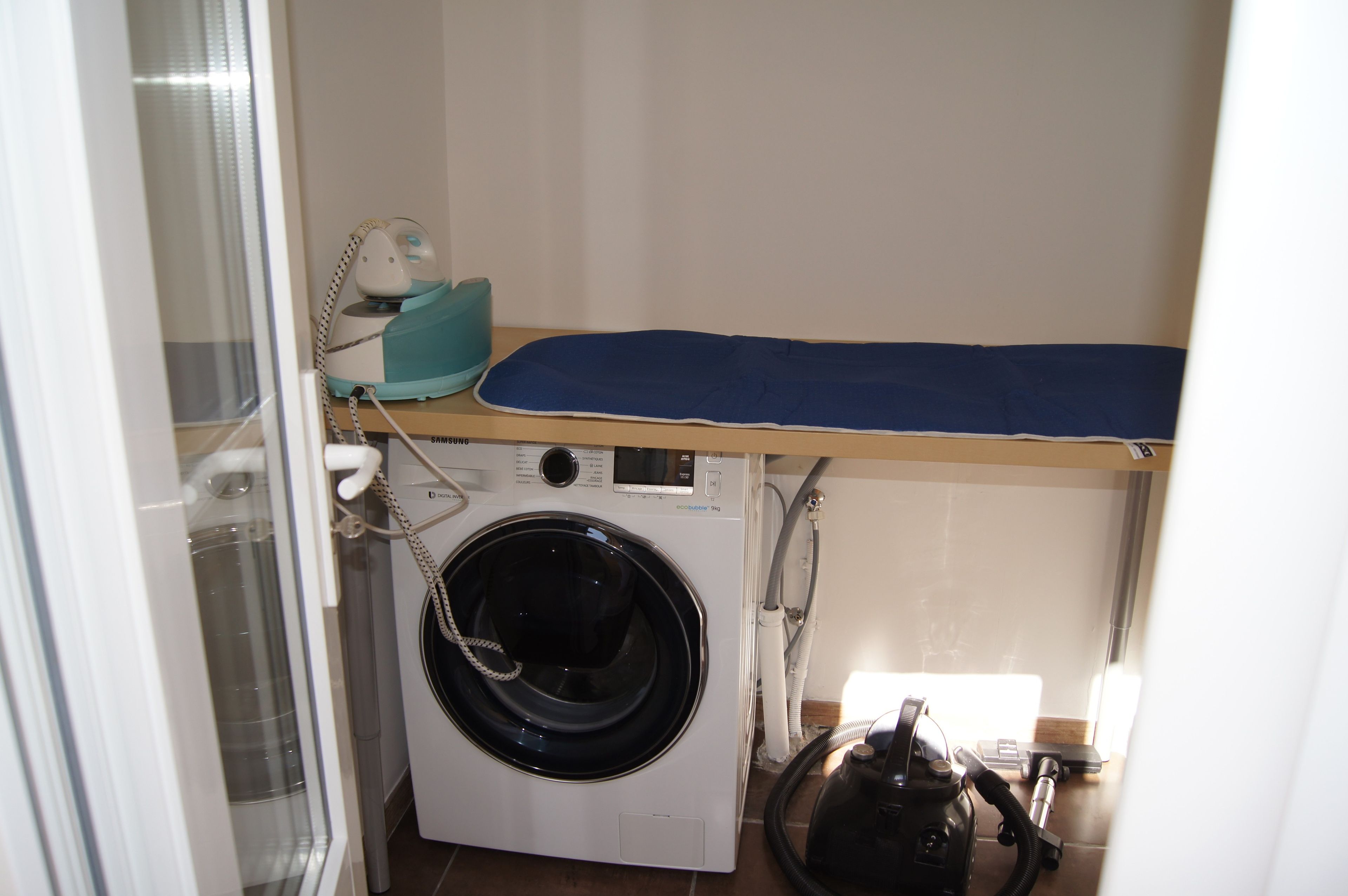 Un espace réduit dédié à la buanderie, comprenant un lave-linge, un étendoir et un aspirateur.