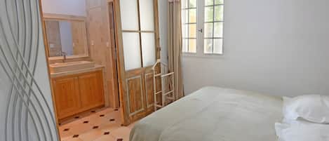 Un dormitorio con cama, ventana, puerta y baño con espejo y lavamanos.